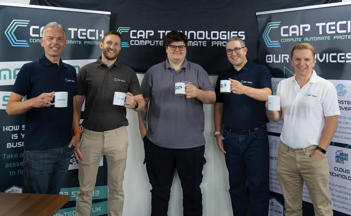The CAP Technologies team together in the Exeter office, holding CAP Tech mugs in front of the company banners
